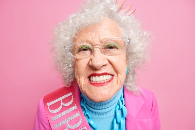 Close up shot of senior grey haired European woman smiles broadly applies bright makeup shows her perfect whit teeth celebrates birthday