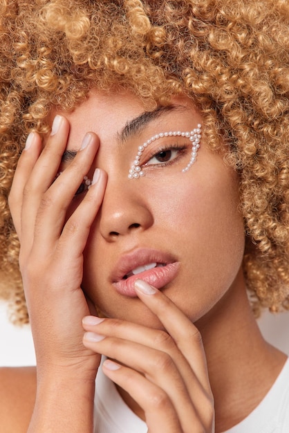 Close up shot of serious woman covers face with hand wears white stones over eye has natural beauty and healthy smooth skin looks confidently at camera has curly hair. Well cared complexion.
