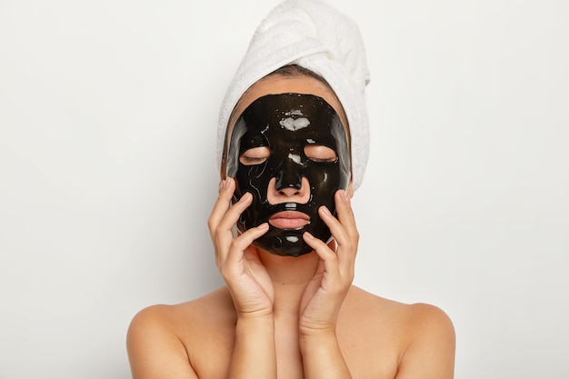 Close up shot of serious young woman wears black facial mask, has eyes closed, touches face gently, wears wrapped towel around head