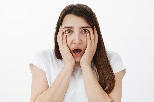 Close-up shot of shocked and speechless concerned young girl hearing shocking rumor gasping dropping jaw from unbelievable news holding hands on cheeks widen eyes impressed