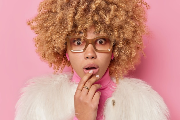 Free photo close up shot of shocked stunned beautiful young woman gasps emotionally holds breath witnesses disaster keeps mouth opened wears spectacles fur coat poses against pink background. omg concept