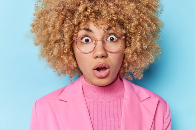 Close up shot of shocked young woman with curly hair keeps mouth opened cannot believe in amazing news finds out amazing relevation wears pink jacket isolated over blue background Omg concept