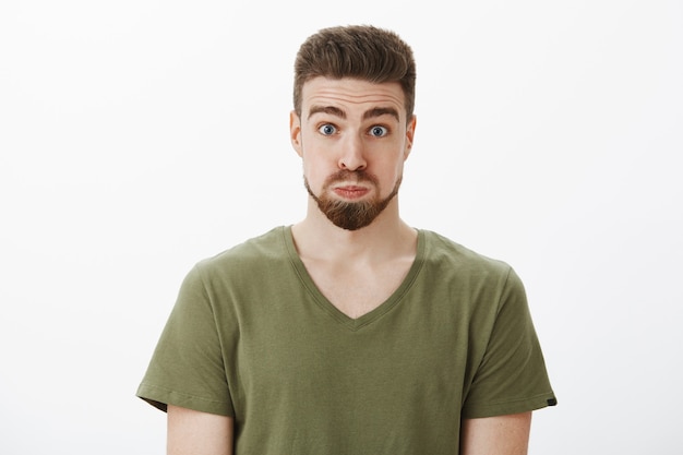 Close-up shot of silly and funny good-looking bearded male in olive t-shirt pouting holding breath looking like balloon raising eyebrows, feeling playful fooling around over white wall