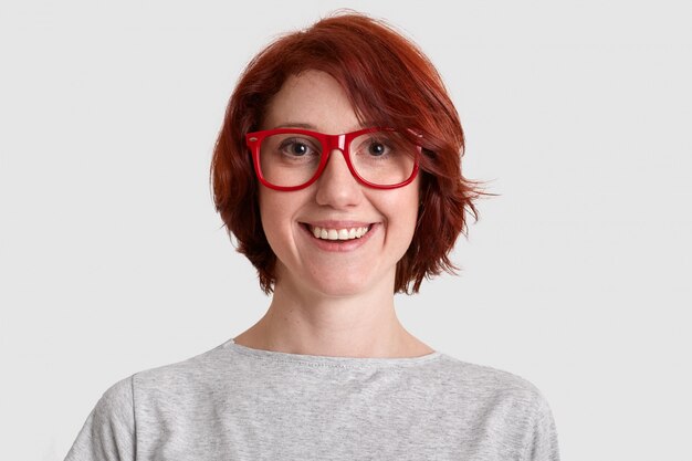 Close up shot of smiling glad woman with short hairstyle, wears red rimed spectacles, dressed casually, isolated over white wall, expresses positive feelings. People and beauty concept.