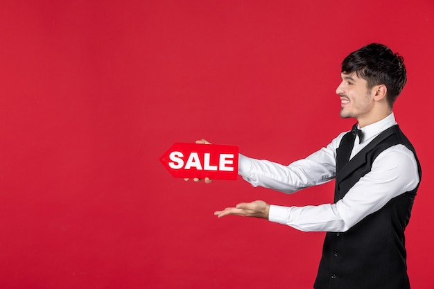 Free photo close up shot of smiling guy waiter in a uniform with butterfly on neck showing sale icon pointing something on the right side on isolated red background