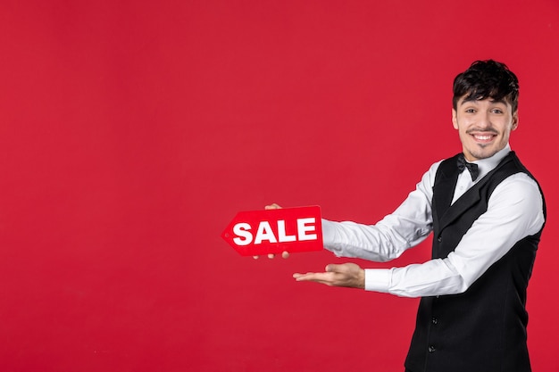 Close up shot of smiling happy guy waiter in a uniform with butterfly on neck showing sale icon pointing something on the right side on isolated red background