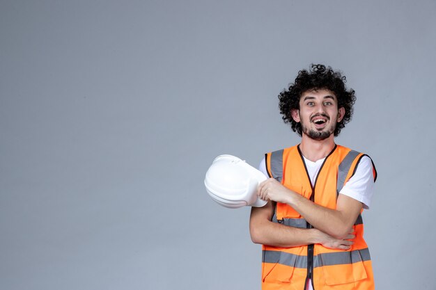 Close up shot of smiling male constructor in warning vest holding safety helmet over gray wave wall