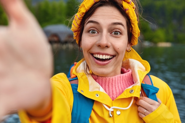 Close up shot of smiling woman stretches hand for making selfie wears yellow headband and raincoat breathes fresh air, stands against river