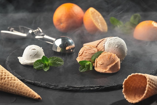 Close-up shot of a stone slate tray served with a tasty creamy and orange ice cream set decorated with mint, and waffle cones on a dark table over a black background. Metal scoop is laying nearby. Sum