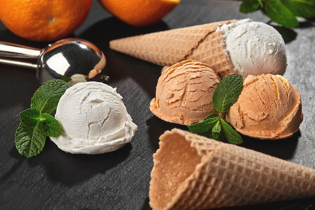 Close-up shot of a stone slate tray with a sweet natural creamy and orange ice cream set decorated with fresh mint, and waffle cones on a dark table over a black background. Metal scoop is laying near