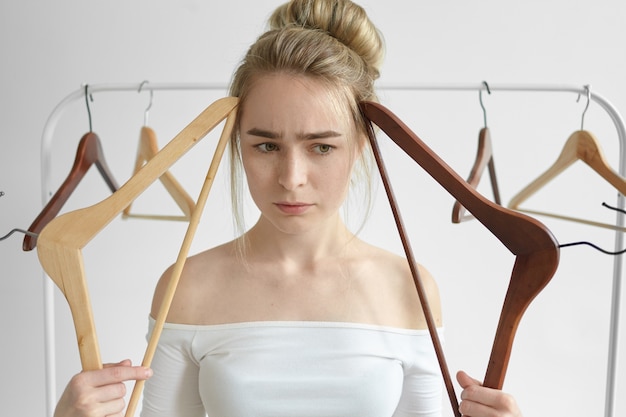 Free photo close up shot of stressed beautiful young female wearing open shoulders white top holding two empty racks at her face and frowning, having perplexed pensive look, thinking what to put on to work