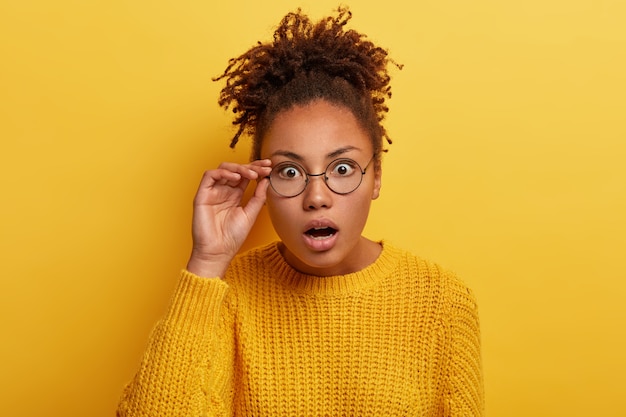 Close up shot of stupefied dark skinned woman reacts to surprising news, keeps hand on rim of eyewear