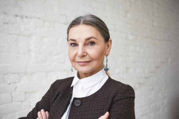 Close up shot of successful beautiful confident senior businesswoman in her fifties with gray hair and blue wise eyes posing indoors, keeping arms folded, looking with charming smile