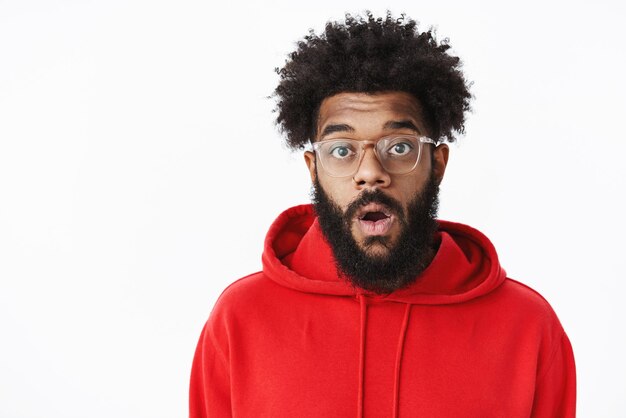 Close-up shot of surprised amazed and speechless african american bearded male with afro hairstyle in glasses and red hoodie dropping jaw from astonishment, being impressed on gray.