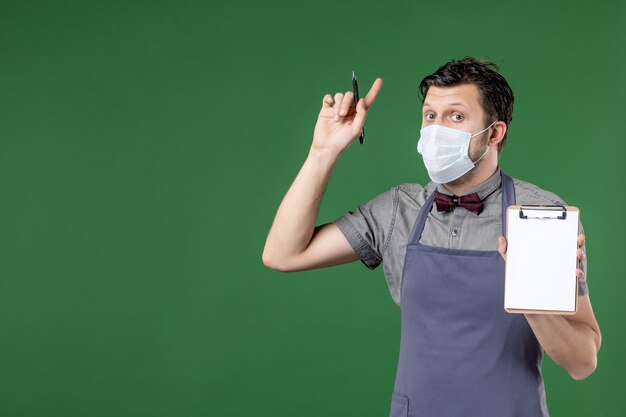 Close up shot of surprised male waiter in uniform with medical mask and holding order book pen pointing up on green background