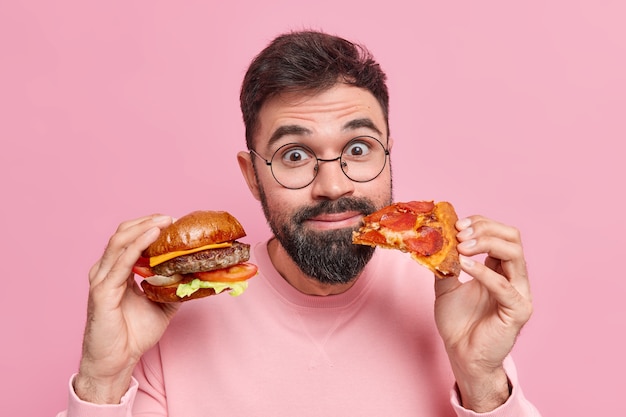 Close up shot of surprised pleased bearded man holds burger and piece of pizza eats junk food doesnt care about health and nutrition wears spectacles neat jumper 