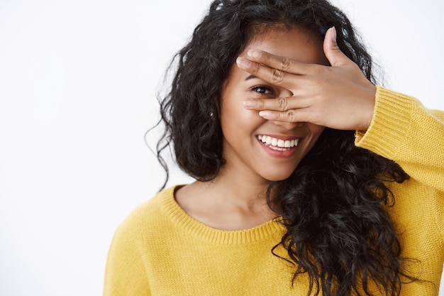 Close-up shot of tender curly-haired girl with toothy smile, holding hand on eye and peeking through fingers, express enthusiasm and joy, standing white wall