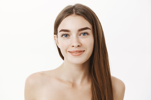Close-up shot of tender and feminine cute young woman with long brown hair standing naked over grey wall and smiling having no pimples being pleased with perfect pure skin condition over grey wall