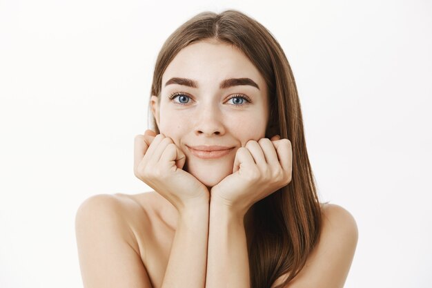 Close-up shot of tender feminine young female brunette posing naked leaning face on palms and smiling gazing charmed with admiration feeling dreamy