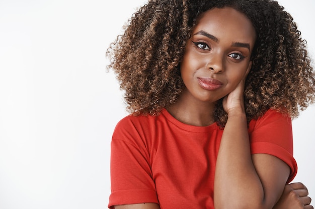 Close-up shot of tender and gentle romantic african-american womanfriend in red casual t-shirt touching back of neck shy and cute tilting head, smiling sensually with flirty gaze over white wall