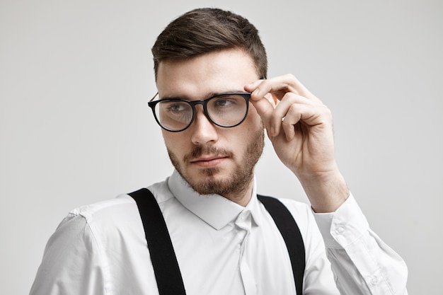 Free photo close up shot of trendy looking hipster guy with trimmed mustache and stubble posing isolated at white studio wall, having confident facial expression, touching his stylish black eyeglasses