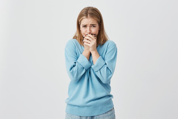 青いセーターとジーンズを着て動揺している10代の少女のショットをクローズアップ。泣きそうになり、顔を隠し、悪い知らせに完全にショックを受けました。