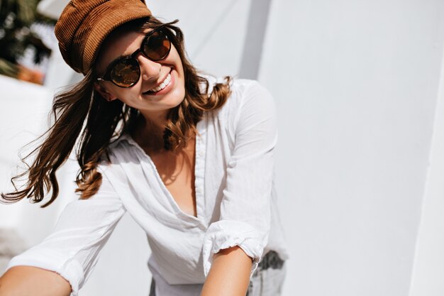 Close-up shot of woman posing on street. Girl in stylish summer outfit and headdress is smiling against space of light house.