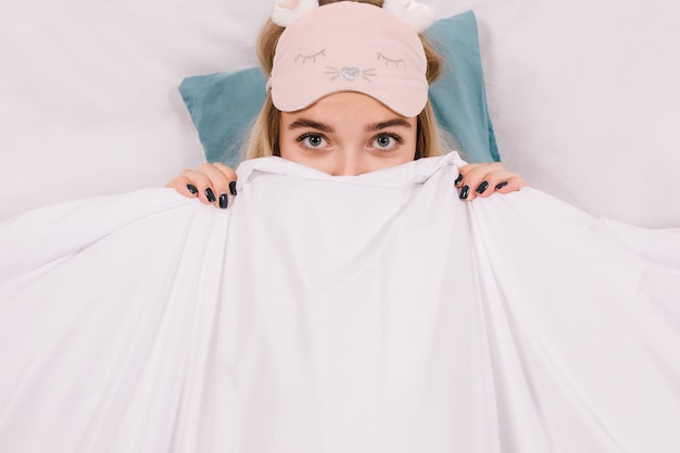 Foto gratuita colpo del primo piano della donna nella maschera di sonno che si nasconde sotto la coperta