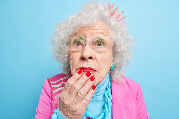 Close up shot of wondered grey haired retired woman keeps hand on chin looks surprisingly