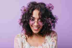 Free photo close-up shot of wonderful female model in stylish sunglasses fooling around. indoor portrait of winsome african girl with purple flowers in black hair.