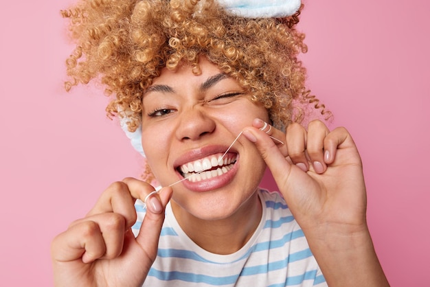 Close up shot of young cheerful woman uses tooth floss for cleaning teeth winks eyes has curly fair hair isolated over pink studio background Health care dental hygiene and stomatology concept