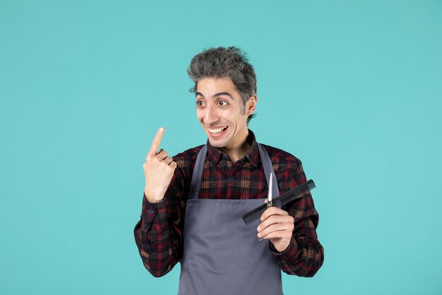 Close up shot of young curious happy male hairdesser wearing gray apron and holding scissor comb pointing up on pastel blue color surface