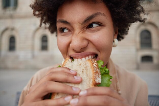 Close up shot of young curly woman feels hungry eats sandwich with appetite poses at street focused somewhere stands outdoors against blurred background prefers junk food. Nutrition concept.