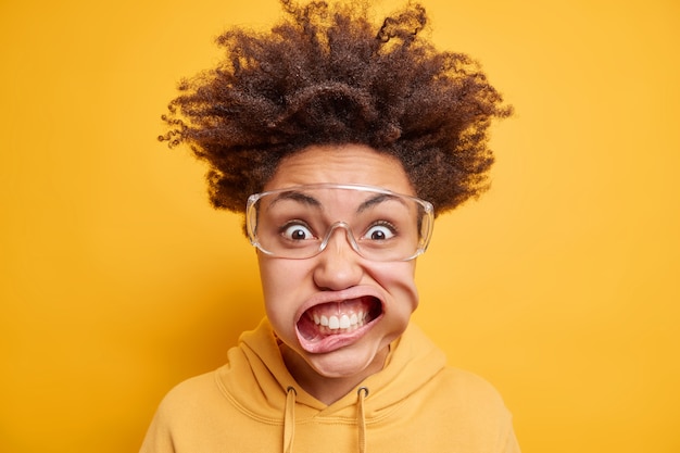 Close up shot of young irritated Afro American woman stares bugged eyes through transparent glasses keeps mouth opened dressed in sweatshirt 