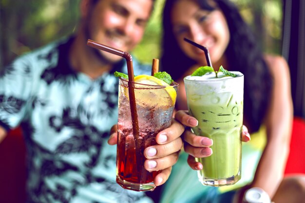 Close up shot of young smiling couple enjoy their drinks, making cheers to camera, matcha latte and berry lemonade, cocktails on party, toned bright colors.