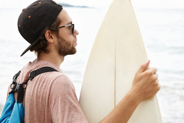 Close up shot of young surfer with stylish beard wearing backpack and baseball cap holding white surfboard, standing on beach and looking at ocean