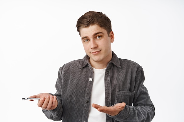 Close up of shrugging boy holding smartphone, looking confused or clueless at camera while using mobile phone app, cant understand something on cellphone, white background
