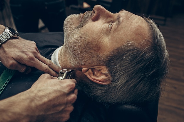 Close-up side back view handsome senior bearded caucasian man getting beard grooming in modern barbershop.