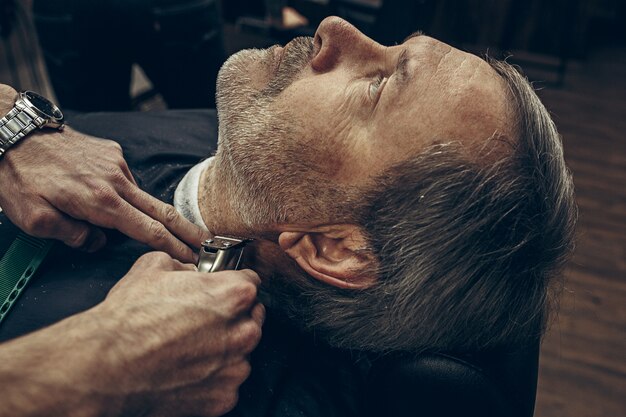 Close-up side back view handsome senior bearded caucasian man getting beard grooming in modern barbershop.