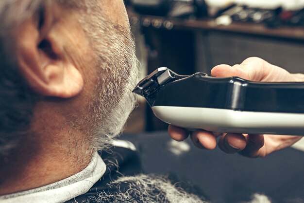 Close-up side back view handsome senior bearded caucasian man getting beard grooming in modern barbershop.