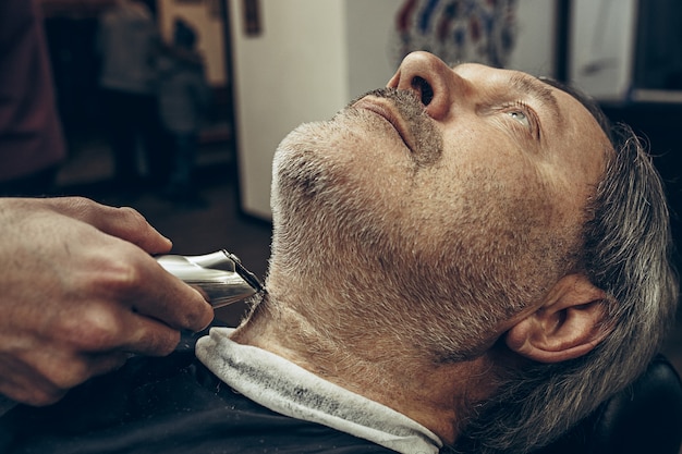 Ritratto laterale di vista di profilo del primo piano dell'uomo caucasico barbuto senior bello che ottiene governare della barba nel parrucchiere moderno.