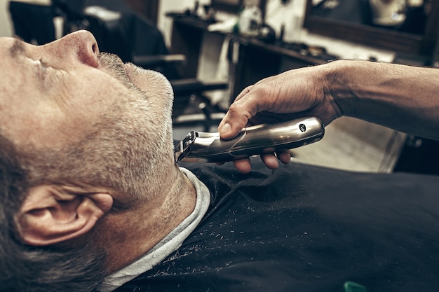 Close-up side profile view portrait of handsome senior bearded caucasian man getting beard grooming in modern barbershop.