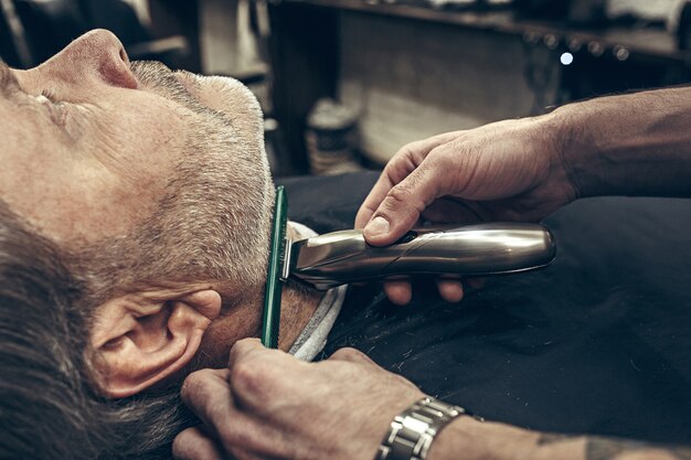 モダンな理髪店でひげの手入れをしているハンサムなシニアのひげを生やした白人男性のクローズアップ側縦断ビューの肖像画。