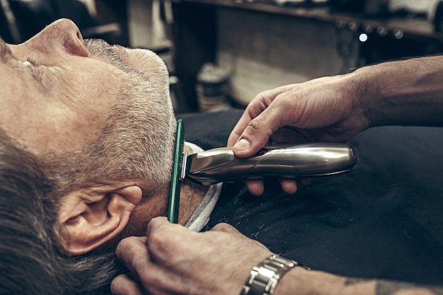 Close-up side profile view portrait of handsome senior bearded caucasian man getting beard grooming in modern barbershop.