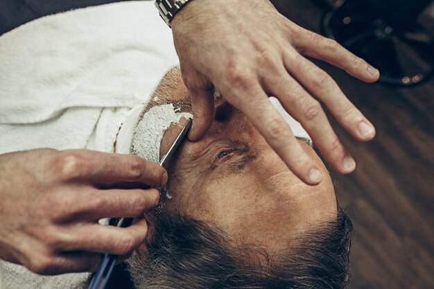 Взгляд сверху стороны конца-вверх красивый старший бородатый кавказский человек получая холить бороды в современной парикмахерскае. Парикмахер обслуживает клиента, делает стрижку бороды прямой бритвой. Концепция парикмахерской.