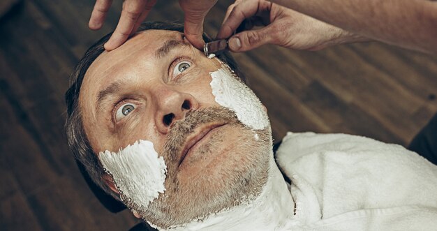 Close-up side top view handsome senior bearded caucasian man getting beard grooming in modern barbershop.