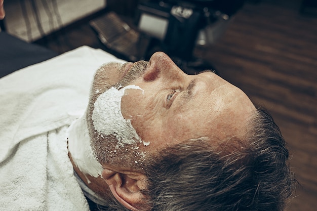 Close-up side top view handsome senior bearded caucasian man getting beard grooming in modern barbershop.