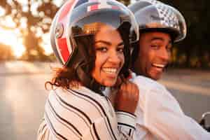 Foto gratuita chiuda sull'immagine di vista laterale delle coppie africane felici che guidano sulla motocicletta moderna all'aperto e che esaminano la macchina fotografica