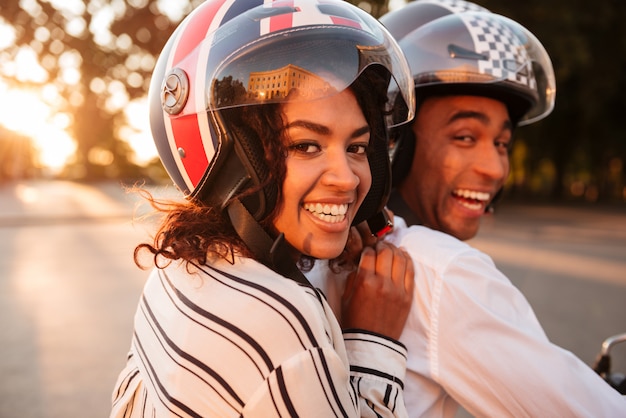 現代のバイクを屋外でカメラに見て幸せなアフリカのカップルの乗り物の側のビューのイメージを閉じます