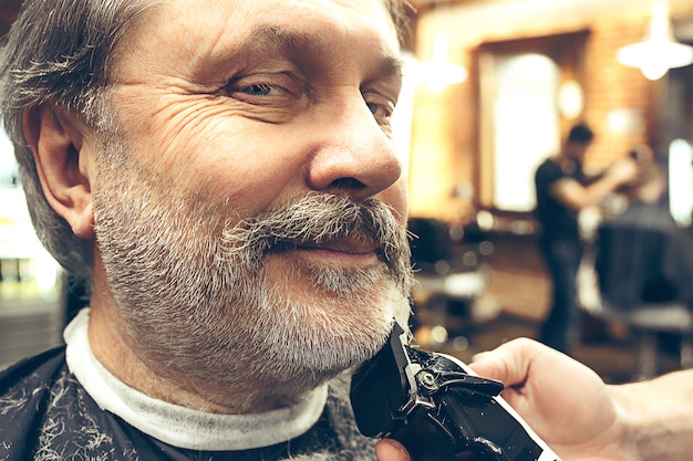 Close-up side view portrait of handsome senior bearded caucasian man getting beard grooming in modern barbershop.